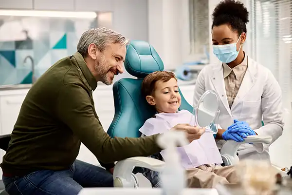 White father holding a mirror for his happy, young son sitting in a dental chair while his Black female dentist happily watches