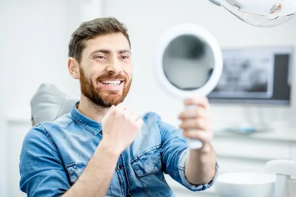 Bearded white patient checking out his handsome smile in a mirror while sitting in a dental chair